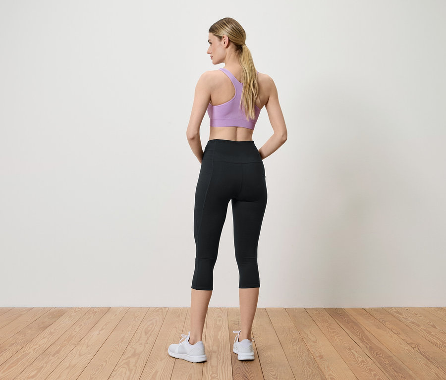 Kobieta pozuje tyłem do kamery, ubrana w fioletowy biustonosz sportowy, czarne legginsy i białe trampki.