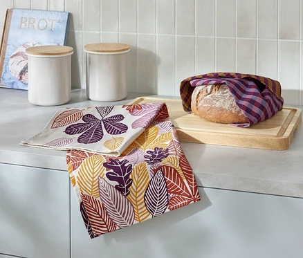 Na blacie kuchennym leży bochenek chleba owinięty w kratkę na drewnianej desce do krojenia, obok dwie ściereczki z nadrukiem liści, dwa białe pojemniki i książka z napisem Brot.