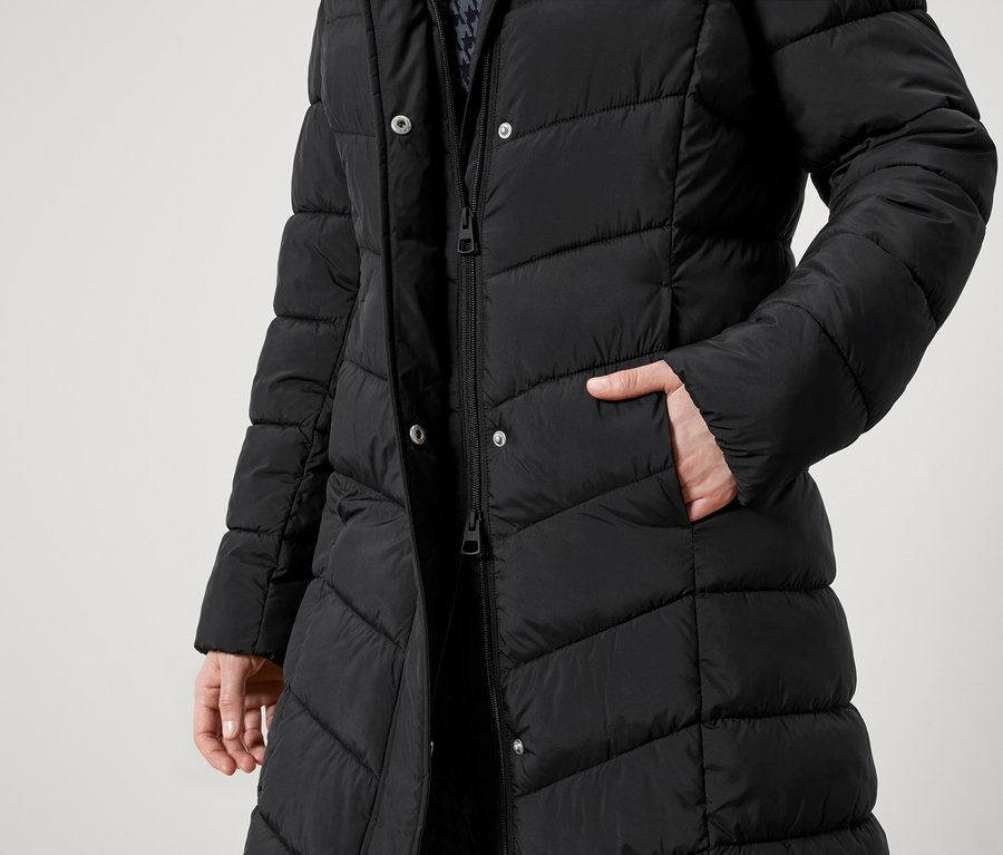 Częściowy widok osoby w czarnym, pikowanym płaszczu z zamkiem błyskawicznym i guzikami. Jedna ręka w kieszeni.