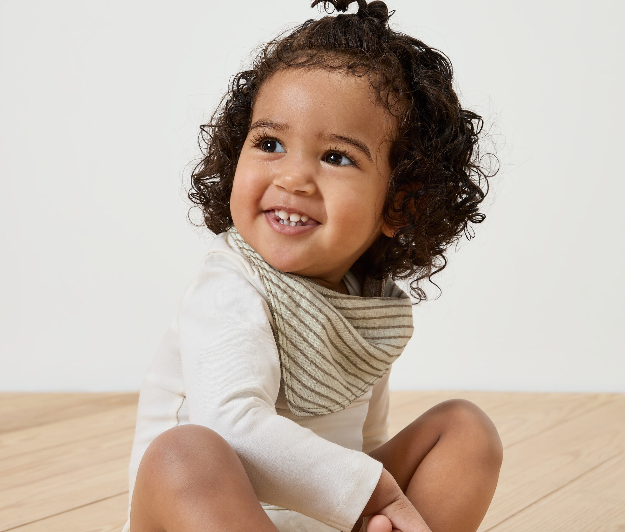 Uśmiechnięte dziecko siedzi na drewnianej podłodze, ma na sobie muślinową chustkę na szyję, 5 sztuk.