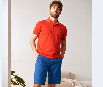 Mężczyzna pozuje w czerwonej koszulce polo i niebieskich szortach chino.