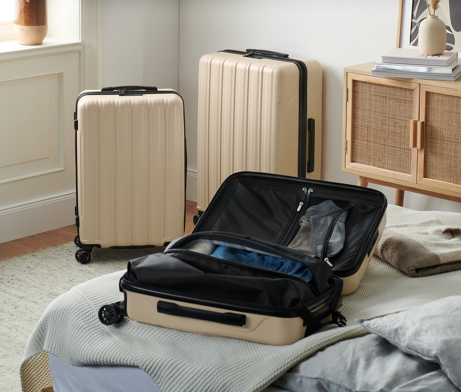 Trzy beżowe walizki z recyklingu stoją w pokoju, jedna otwarta na łóżku.
