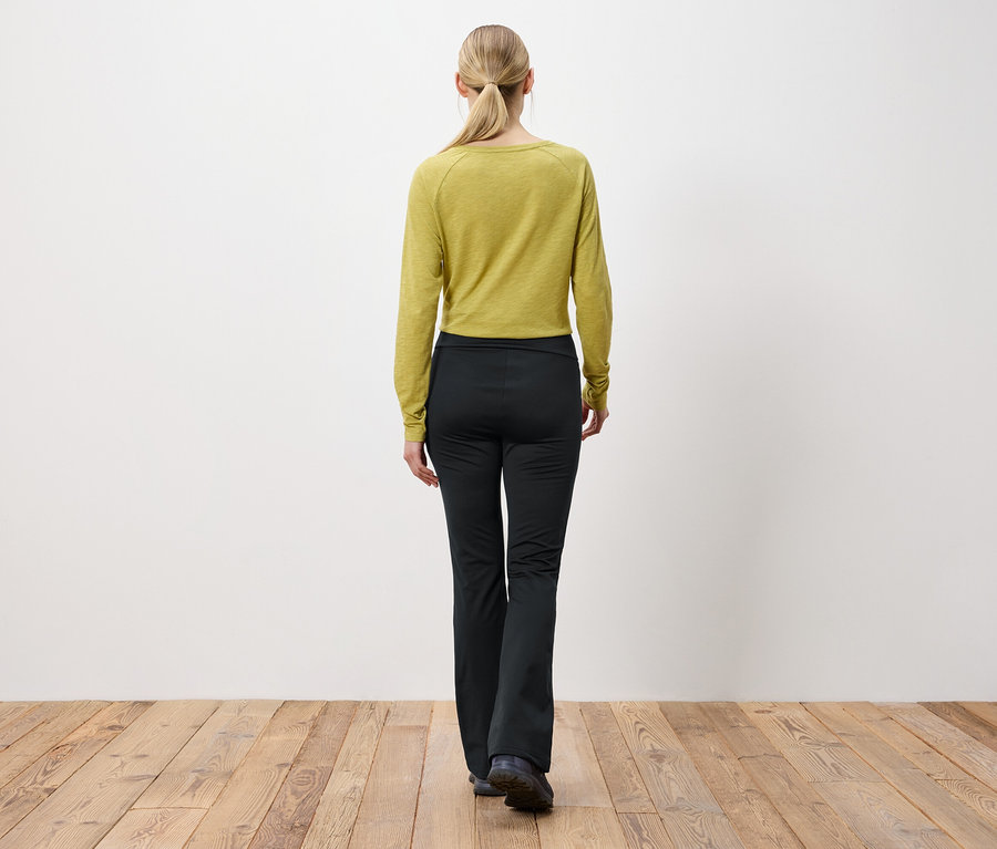 Kobieta o blond włosach związanych w kucyk stoi tyłem do kamery, ubrana w oliwkowozieloną bluzkę z długim rękawem i czarne spodnie.
