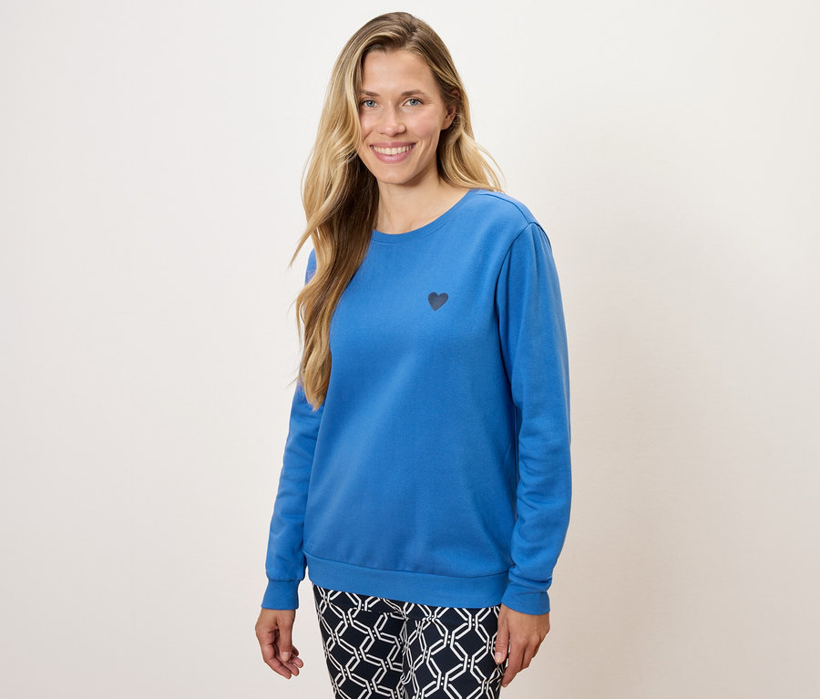 Kobieta ma na sobie niebieską bluzę z długim rękawem z małym czarnym sercem i wzorzyste spodnie.