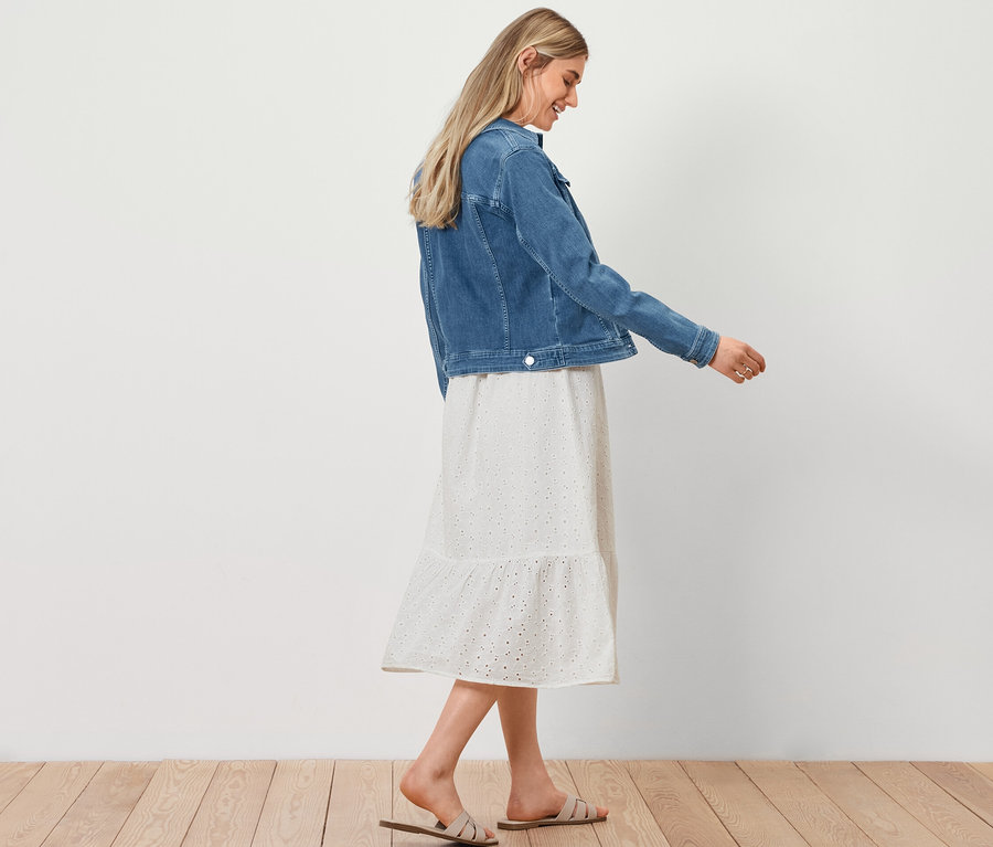 Kobieta idzie, ma na sobie kurtkę dżinsową, sukienkę koszulową z haftem ażurowym i torbę shopperkę z plecionki.