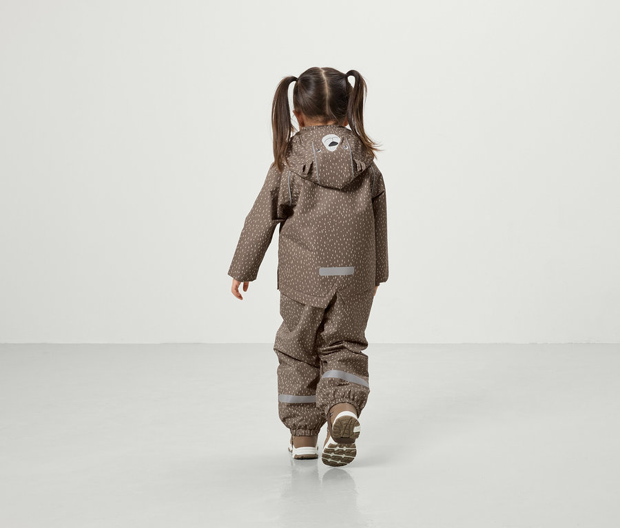 Dziewczynka z warkoczami idzie po podłodze w brązowej kurtce z kapturem i spodniach.