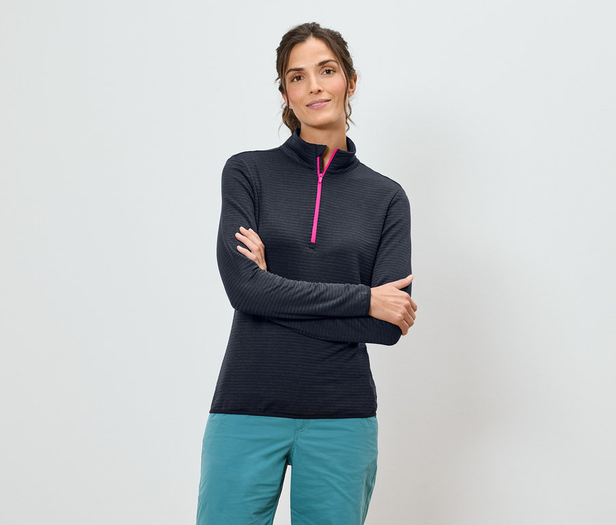 Kobieta ze skrzyżowanymi ramionami ma na sobie granatową koszulę z długim rękawem z różowym zamkiem błyskawicznym i turkusowe spodnie na białym tle.