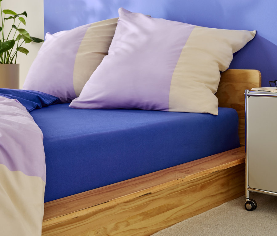 Łóżko z niebieskim prześcieradłem z gumką i poduszkami w fioletowe i beżowe paski.