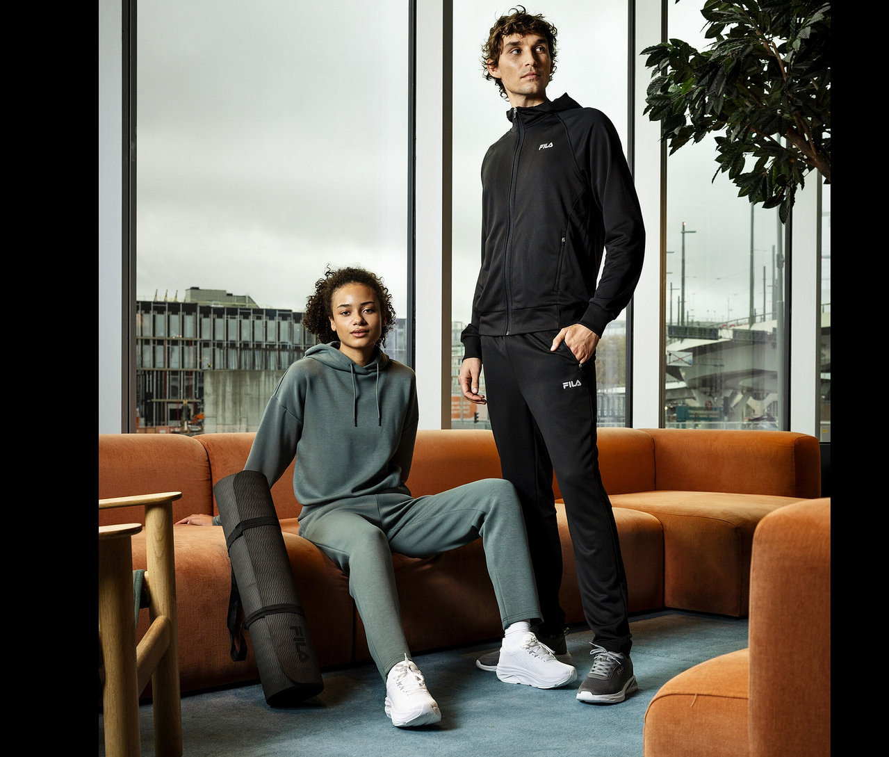 Kobieta siedzi na pomarańczowej sofie z czarną matą do jogi. Obok stoi mężczyzna. Oboje noszą odzież sportową marki Fila.