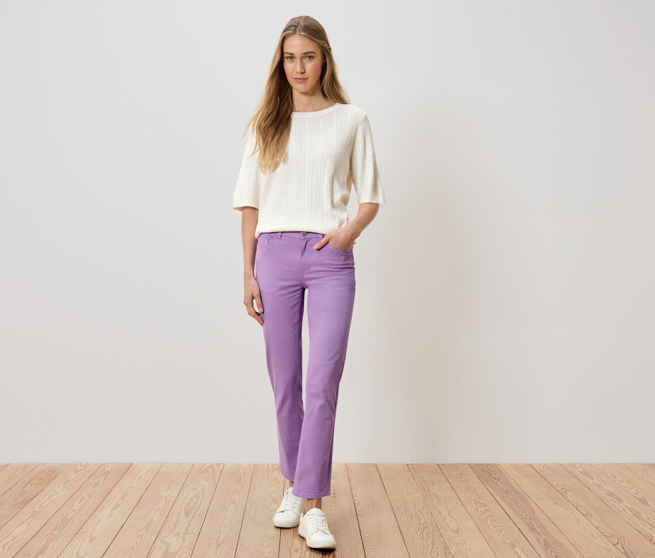 Kobieta stoi z ręką w kieszeni, ubrana w fioletowe spodnie, biały sweter i białe buty.
