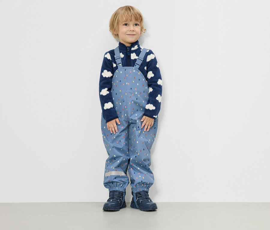 Mały chłopiec stoi na tle białej ściany, ubrany w niebieską bluzę polarową w chmury i niebieskie spodnie przeciwdeszczowe w kolorowe kropki.
