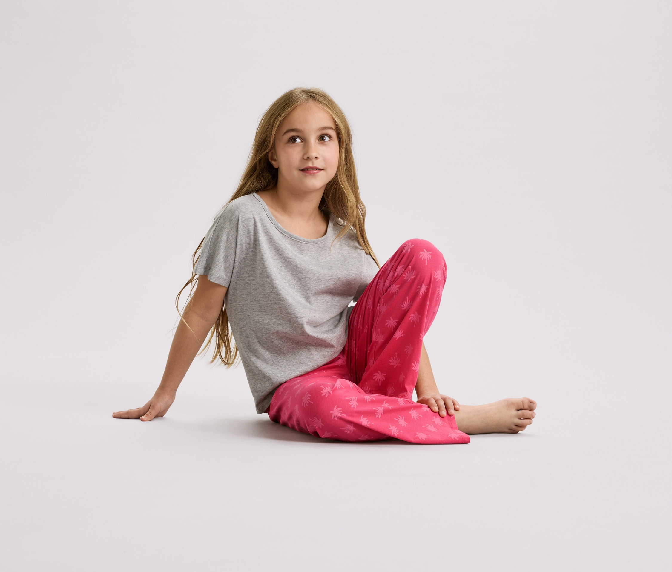 Dziewczynka siedzi na podłodze, ubrana w szary T-shirt i różowe spodnie w palmy.