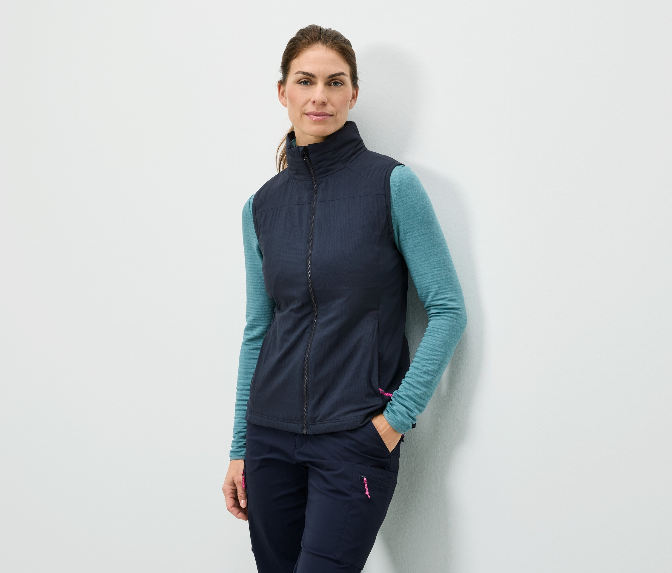 Kobieta stoi przy ścianie z ręką w kieszeni, ubrana w granatową kamizelkę, turkusową bluzkę z długim rękawem i granatowe spodnie.