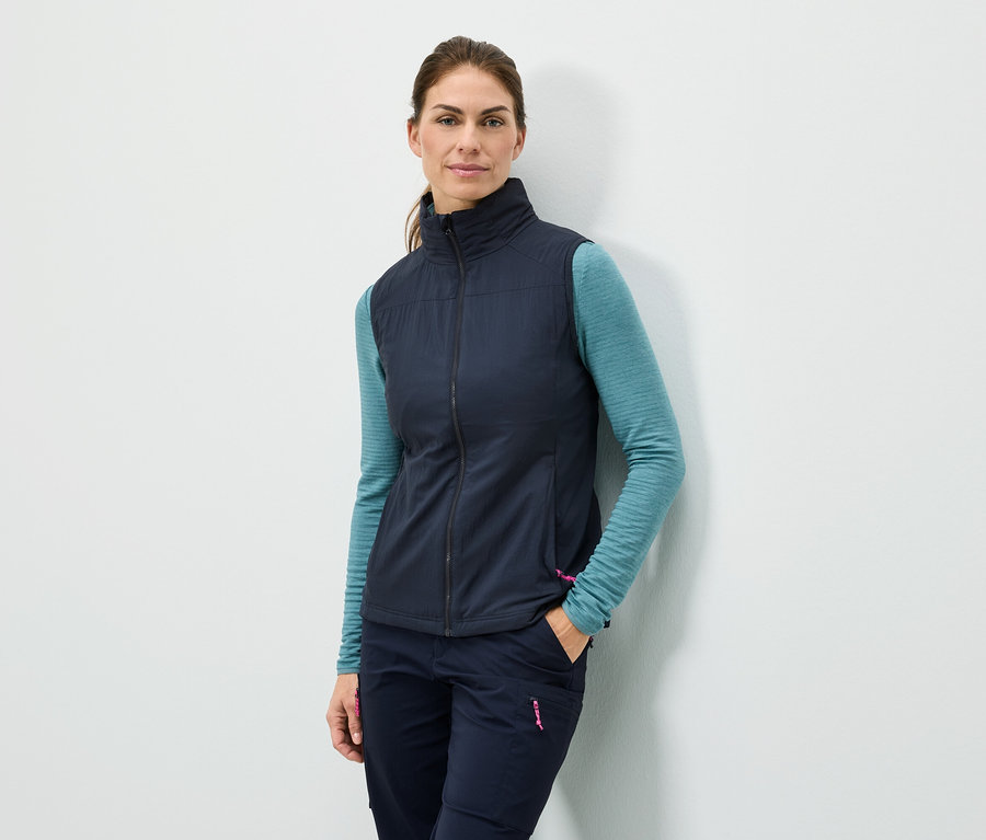 Kobieta stoi przy ścianie z ręką w kieszeni, ubrana w granatową kamizelkę, turkusową bluzkę z długim rękawem i granatowe spodnie.