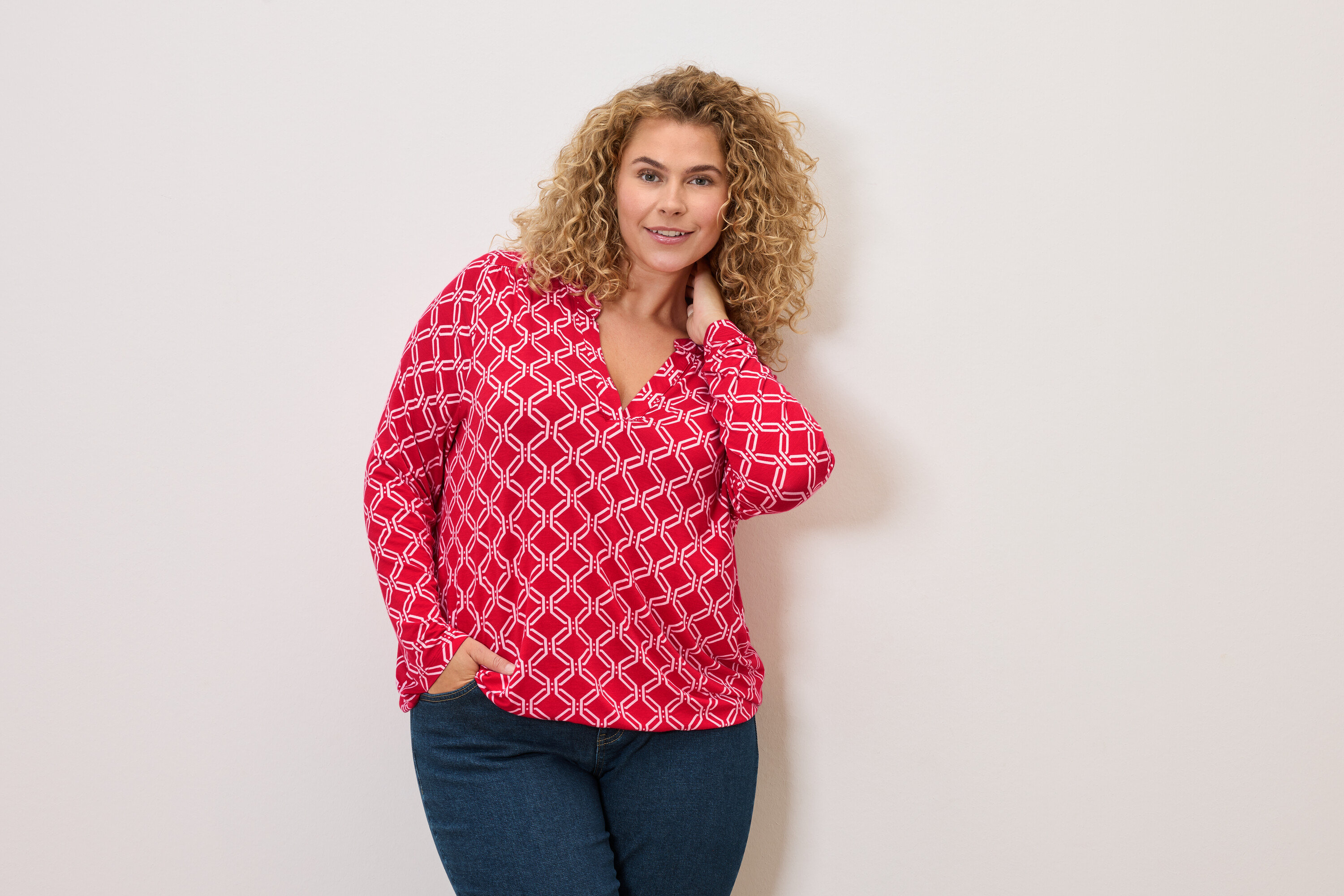 Kobieta o kręconych włosach w czerwono-białej bluzce i dżinsach, pozująca z ręką w kieszeni.