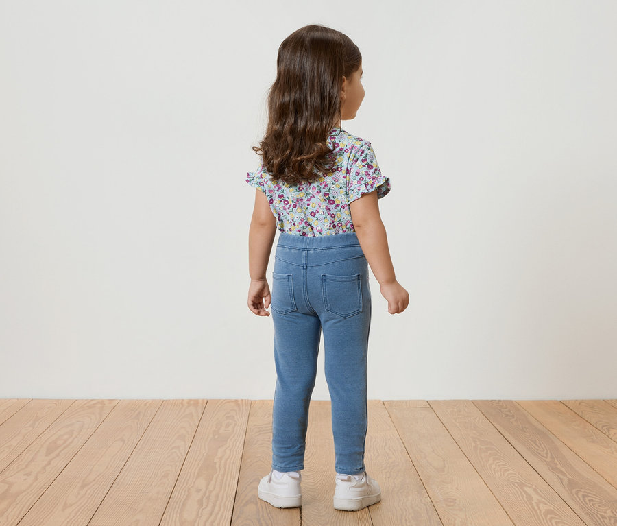 Widok z tyłu małej dziewczynki z kręconymi włosami, ubranej w niebieską bluzkę w kwiaty z krótkim rękawem, niebieskie legginsy i białe tenisówki.