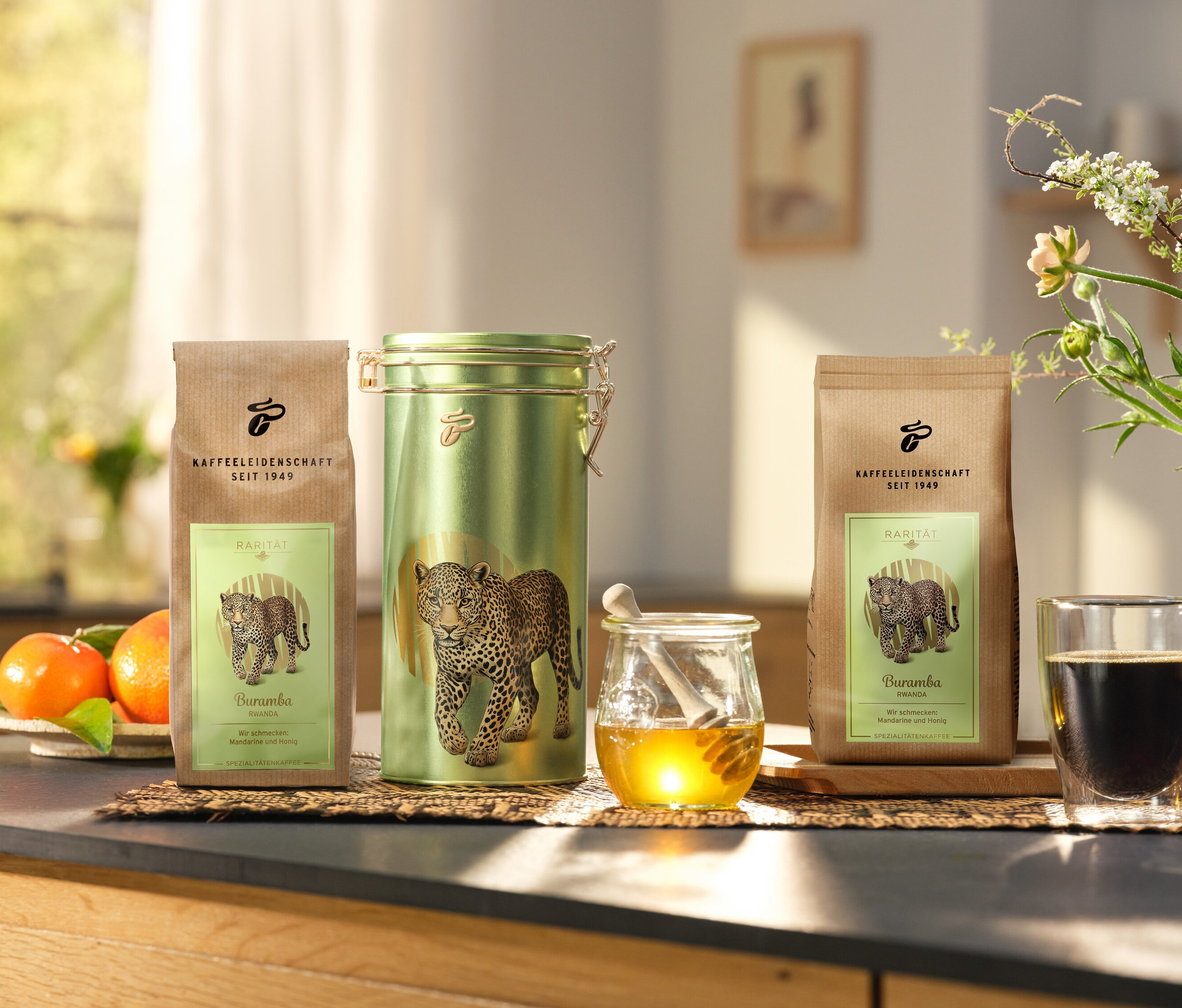 Na blacie kuchennym stoją puszka kawy i dwa opakowania »Buramba Rwanda« wraz z puszką kawy obok filiżanki kawy i miodu.