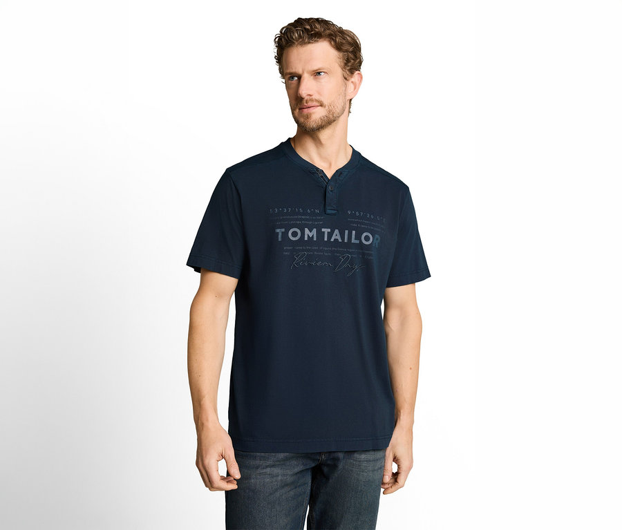 Mężczyzna z kręconymi brązowymi włosami i brodą, ubrany w granatowy T-shirt z krótkim rękawem z dekoltem Henley i napisem "TOM TAILOR" oraz ciemne dżinsy. Stoi na białym tle i patrzy w prawo.