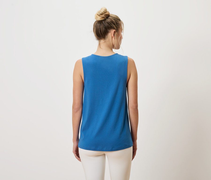 Tył kobiety z kokiem na głowie, ubranej w niebieski top na ramiączkach i białe spodnie.