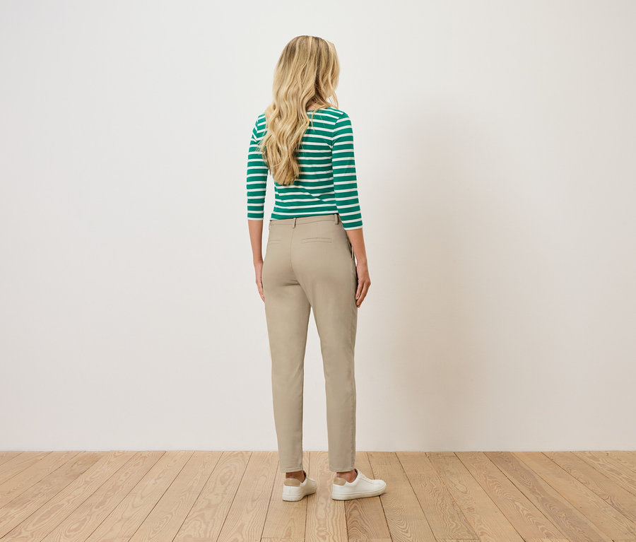 Kobieta o blond, falowanych włosach stoi tyłem do widza, ubrana w bluzkę w zielono-białe paski, beżowe spodnie i białe tenisówki.