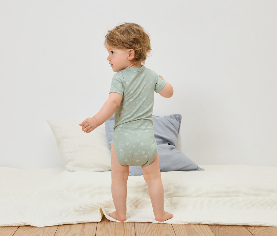 Małe dziecko w jasnozielonym body w serduszka stoi na białym kocyku do zabawy.