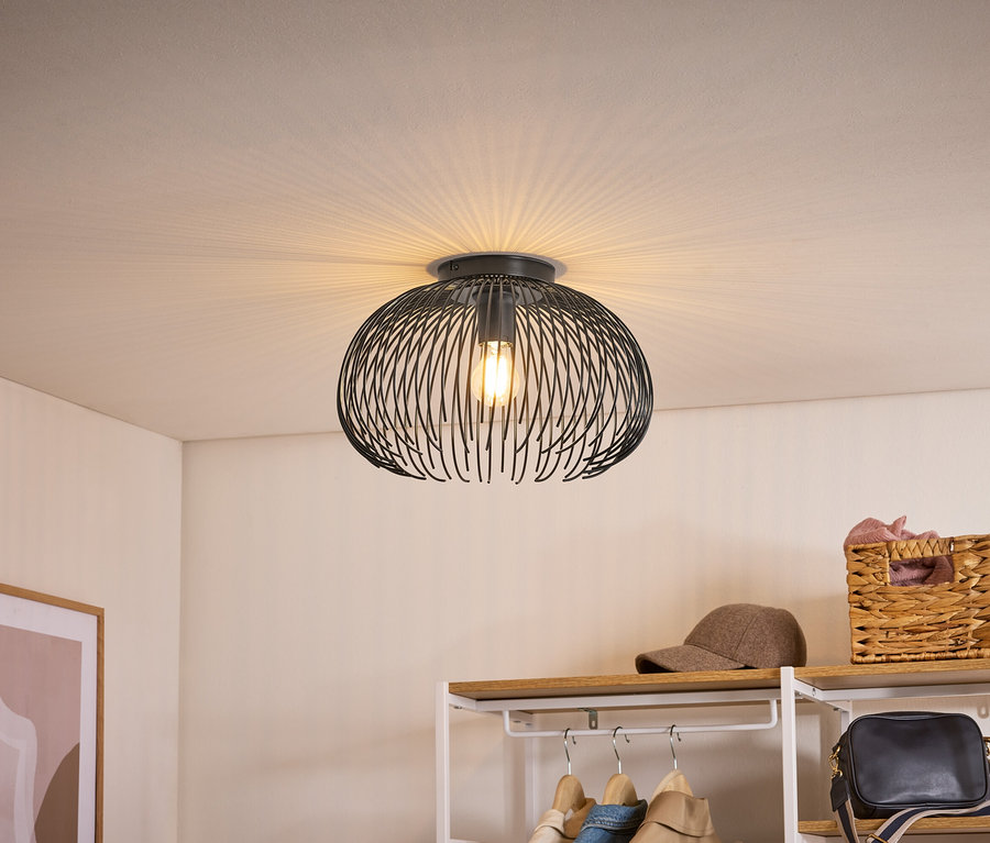 Czarna lampa sufitowa z drucianym kloszem, rzucająca światło w kształcie wachlarza na sufit. Poniżej regał z ubraniami, czapką i koszem.
