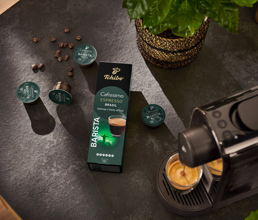 Ekspres do kawy napełnia filiżankę. W tle opakowanie kapsułek BARISTA Espresso Brasil – 10 kapsułek i ziarno kawy.
