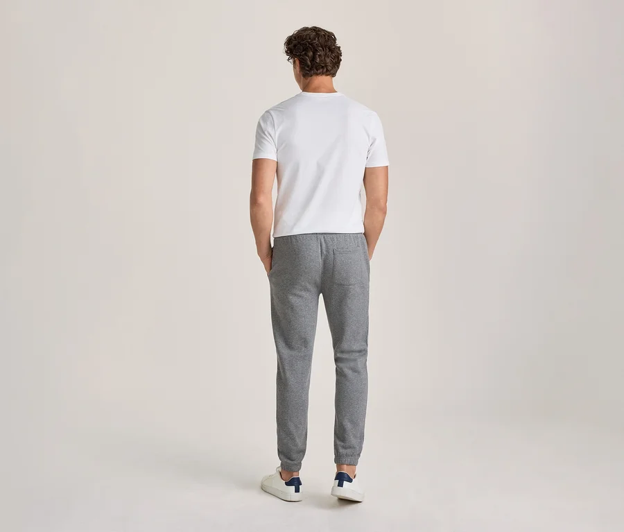 Mężczyzna widziany od tyłu, ubrany w biały T-shirt, szare spodnie dresowe z rękami w kieszeniach i białe sneakersy.