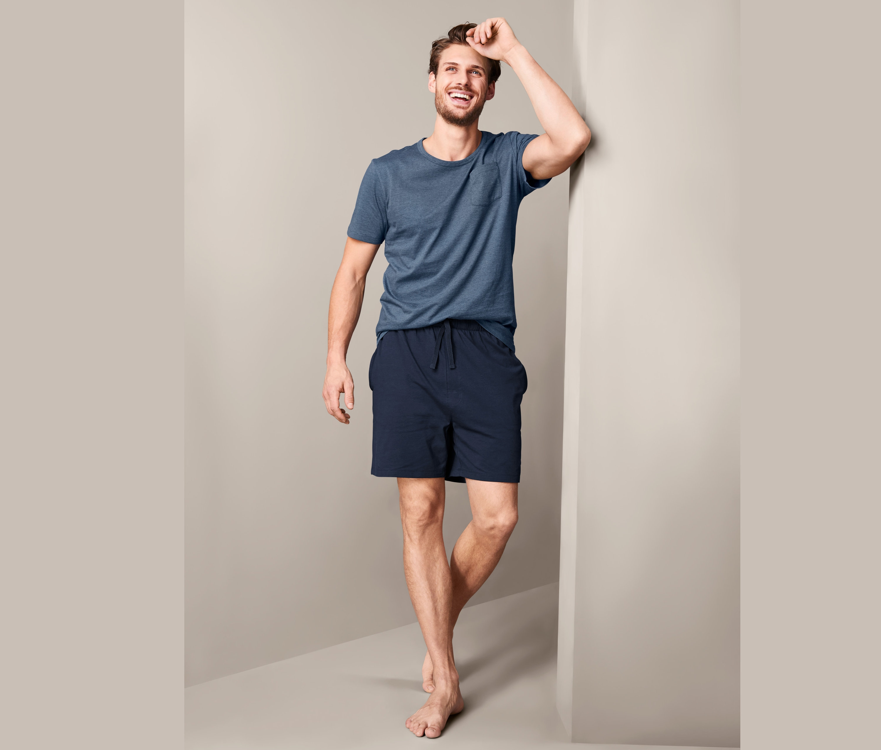 Uśmiechnięty mężczyzna opiera się o ścianę, ubrany w męską piżamę ze spodenkami.