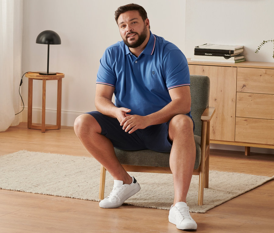 Mężczyzna siedzi w fotelu, ubrany w granatowe szorty chinosy, niebieską koszulkę polo o splocie piqué i buty typu sneaker.