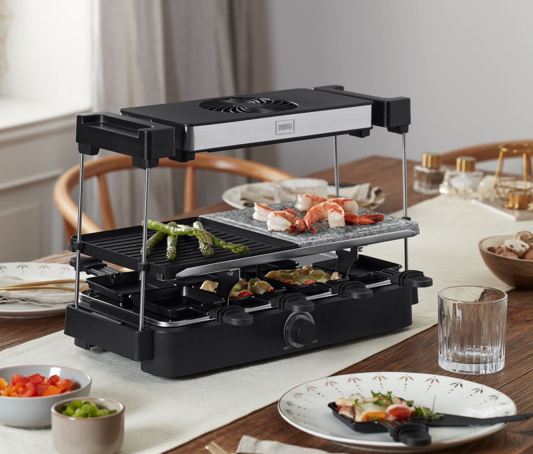 Na drewnianym stole stoi grill raclette Trebs z okapem i płytą kamienną »15100«, na którym grillowane są potrawy.