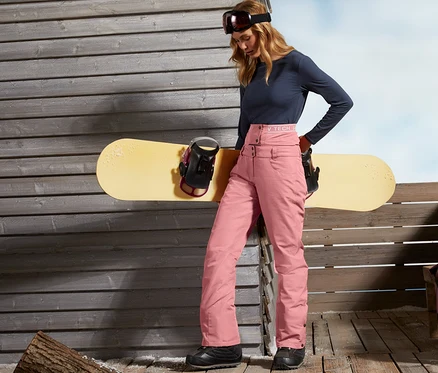 Kobieta stoi przy drewnianej ścianie i opiera się o deskę snowboardową. Ma na sobie spodnie narciarskie i snowboardowe.