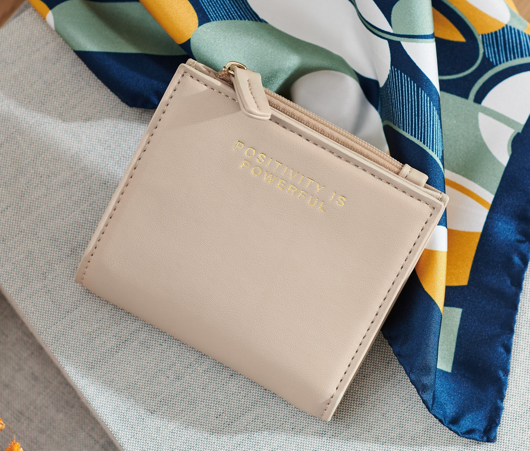 Kremowa portmonetka leży na szarej tkaninie obok kwiatów i szalika.