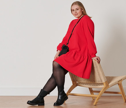 Kobieta siedzi na wiklinowym krześle, ubrana w czerwoną sukienkę z dzianiny dresowej, buty skórzane, rajstopy (2 pary) i torebki mini.