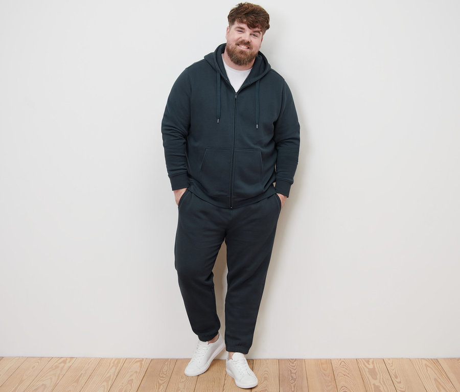 Mężczyzna pozuje do zdjęcia w rozpinanej bluzie z kapturem, spodniach dresowych i białych tenisówkach.