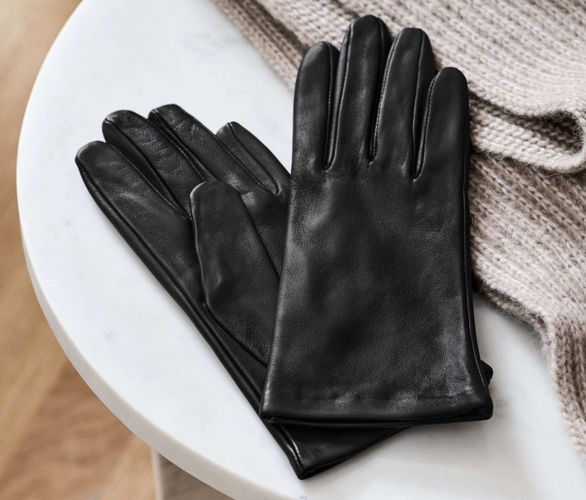 Czarne skórzane rękawiczki leżą na białym marmurowym stoliku obok beżowego swetra.