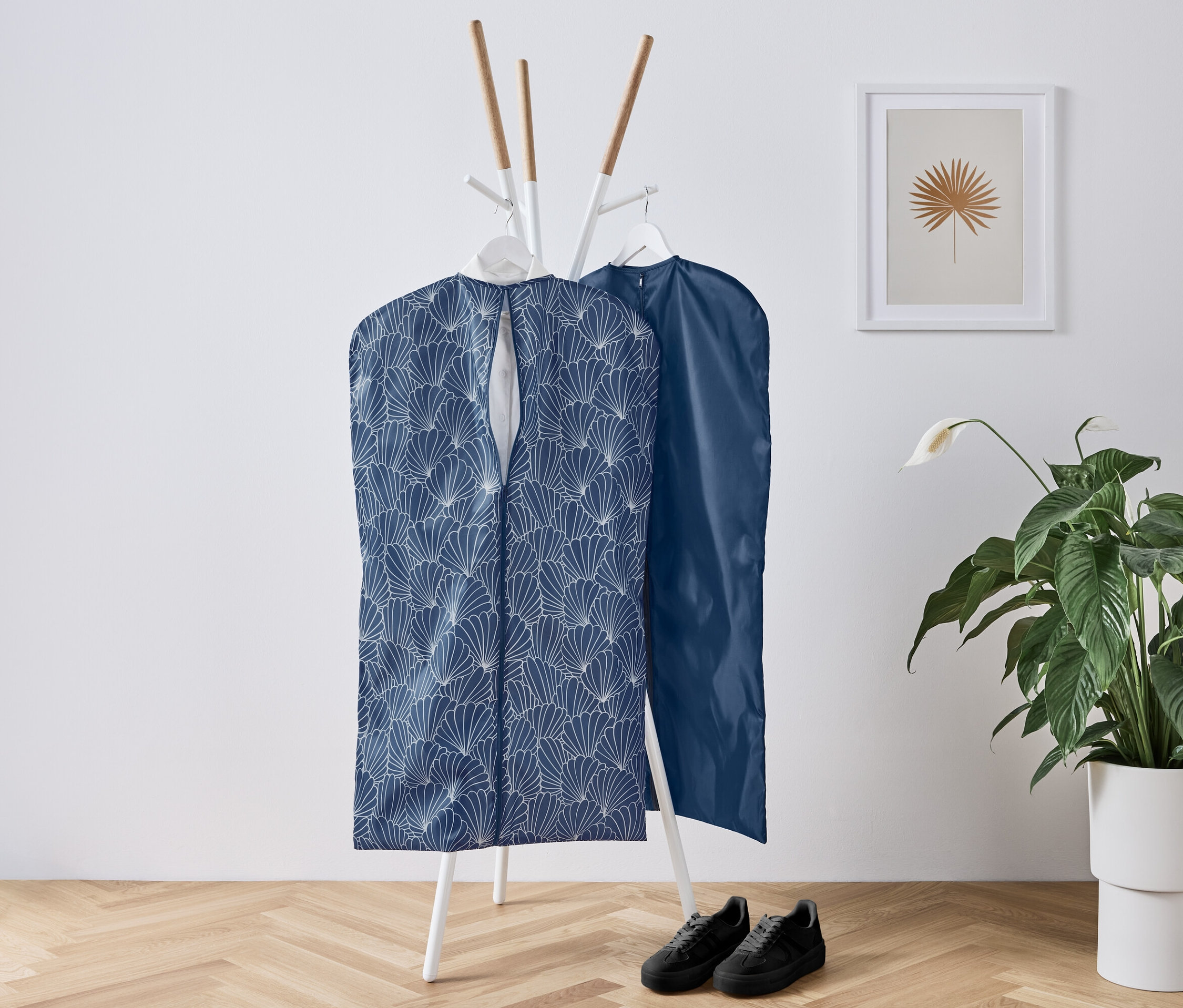 Dwa pokrowce na ubrania wiszą na białym wieszaku, pod nimi stoją czarne buty, obok kwiat.