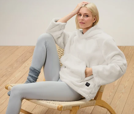 Blondwłosa kobieta siedzi na krześle i ma na sobie ogrzewany sweter z akumulatorem.