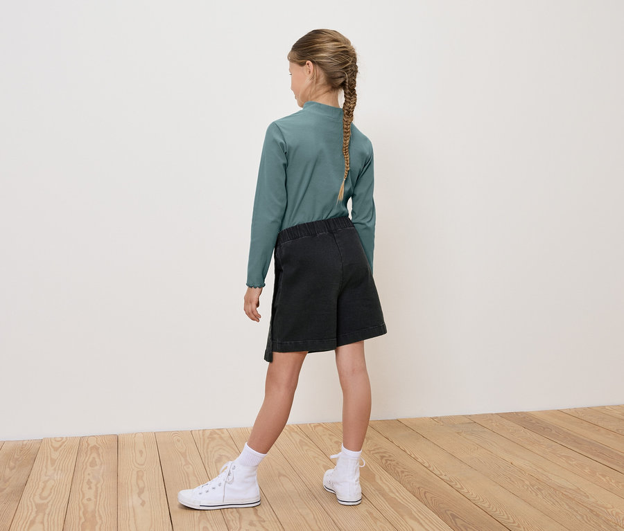 Dziewczynka tyłem do kamery ma na sobie zieloną bluzkę z długim rękawem, czarne szorty i białe tenisówki.