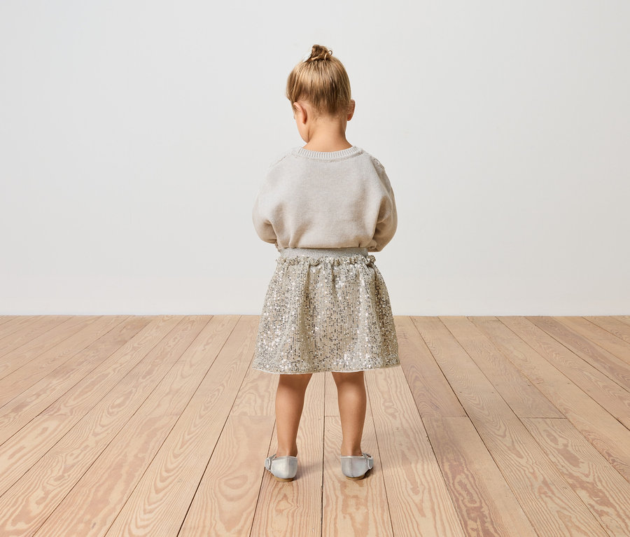 Dziewczynka stoi tyłem, ubrana w brokatową spódnicę i sweter.