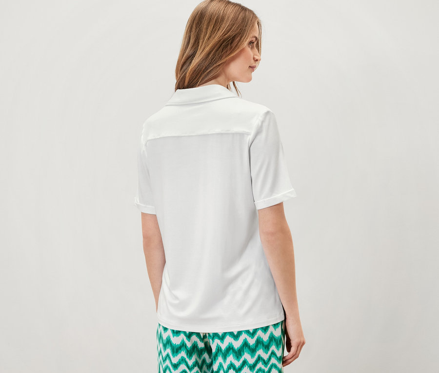 Kobieta stoi tyłem do aparatu, ubrana w białą bluzkę zapinaną na guziki i zielone wzorzyste spodnie.