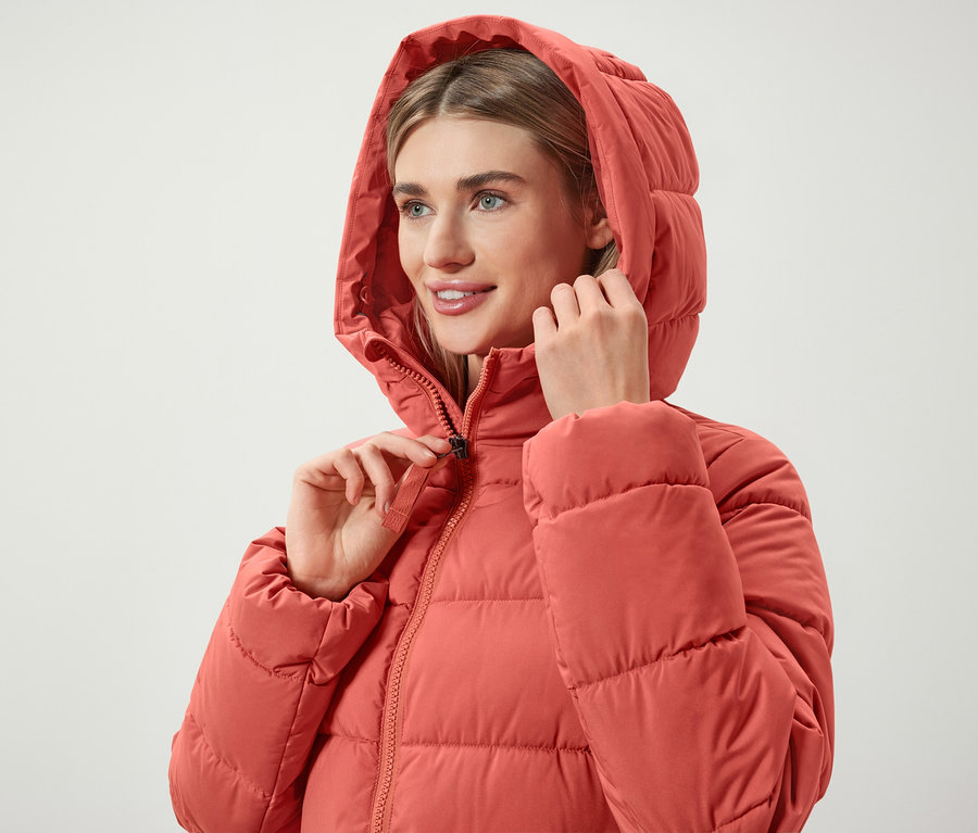 Kobieta zapina zamek błyskawiczny w koralowej puchowej kurtce z kapturem.