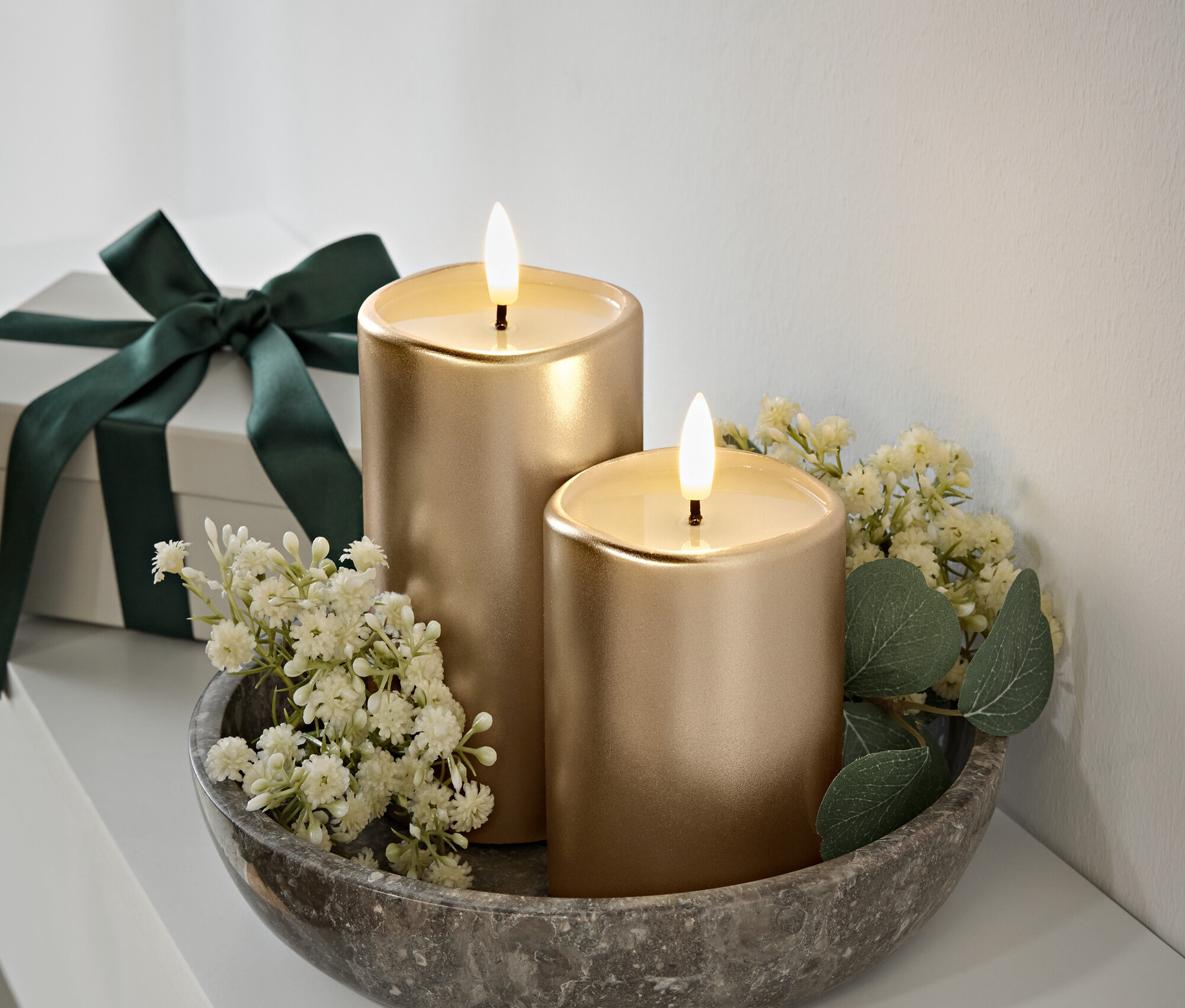Dwie złote świece z płomieniami w szarej kamiennej misce, białe kwiaty i zielone liście. Pudełko prezentowe z ciemnozieloną wstążką w tle.