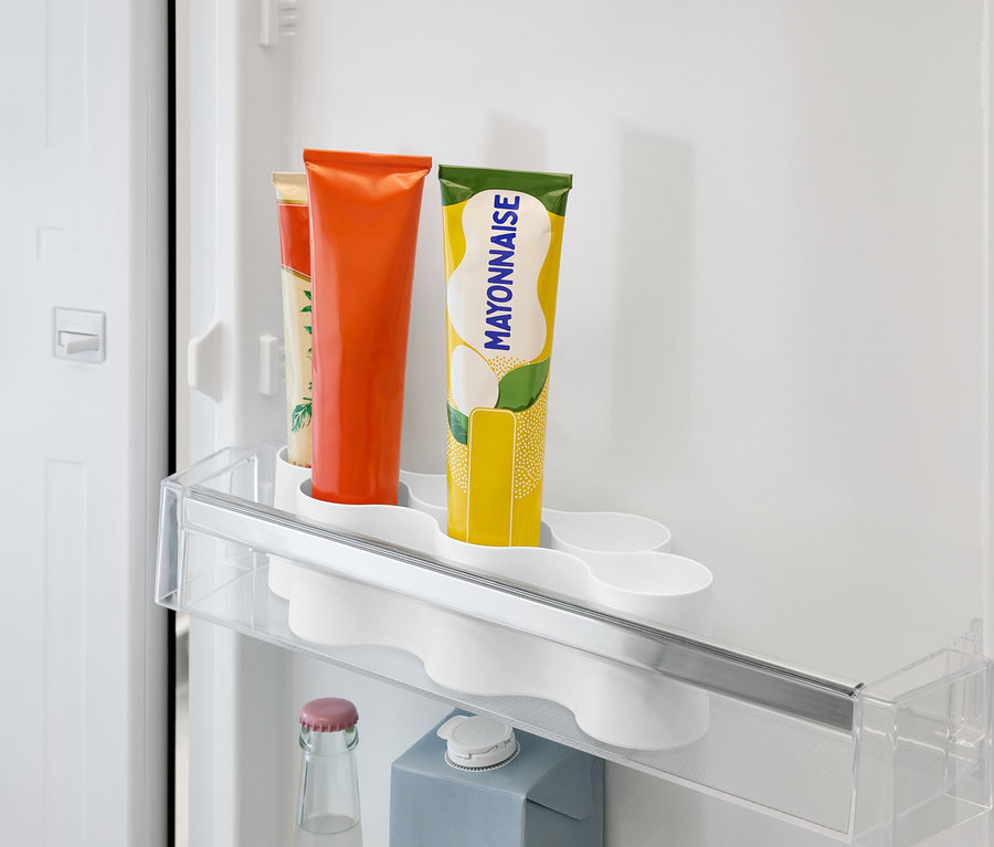 W lodówce znajduje się przezroczysta półka z tubkami z pomarańczowymi, żółtymi i beżowymi etykietami, a pod nią butelka i karton.