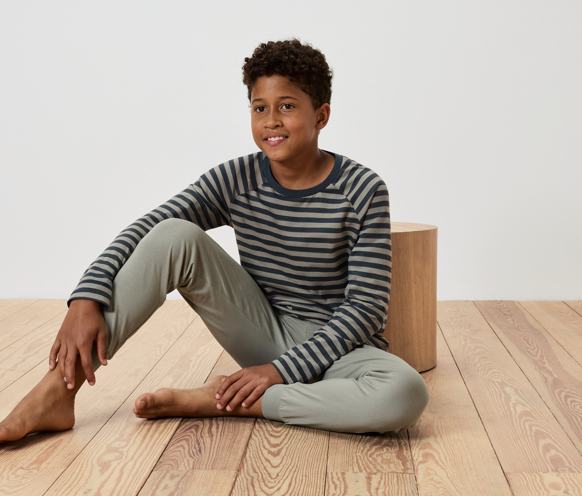 Młody chłopak siedzi na drewnianej podłodze, opierając się o mały drewniany stół, ubrany w koszulkę w paski i spodnie.