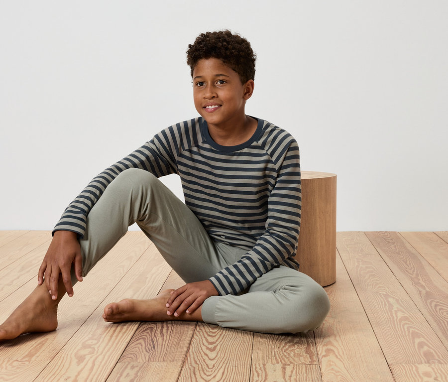 Młody chłopak siedzi na drewnianej podłodze, opierając się o mały drewniany stół, ubrany w koszulkę w paski i spodnie.