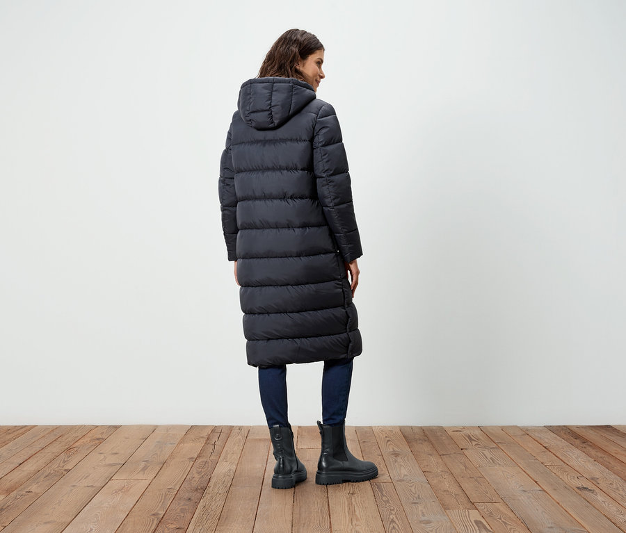 Kobieta tyłem do kamery ma na sobie długi czarny puchowy płaszcz, dżinsy i buty.