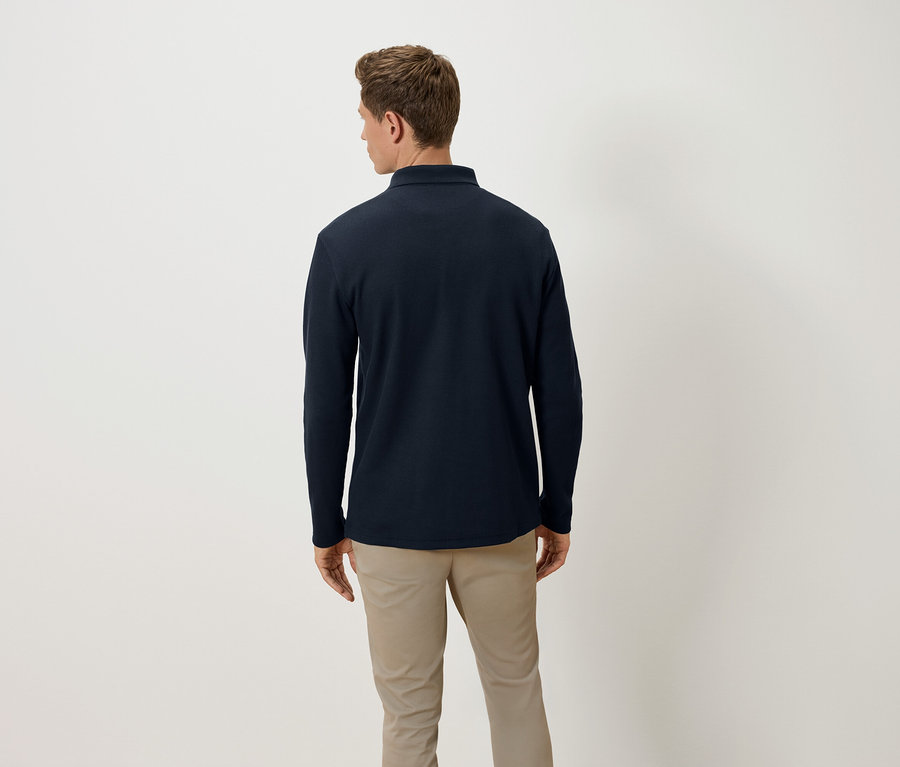 Mężczyzna tyłem do kamery ma na sobie granatową koszulkę polo z długim rękawem i beżowe spodnie.