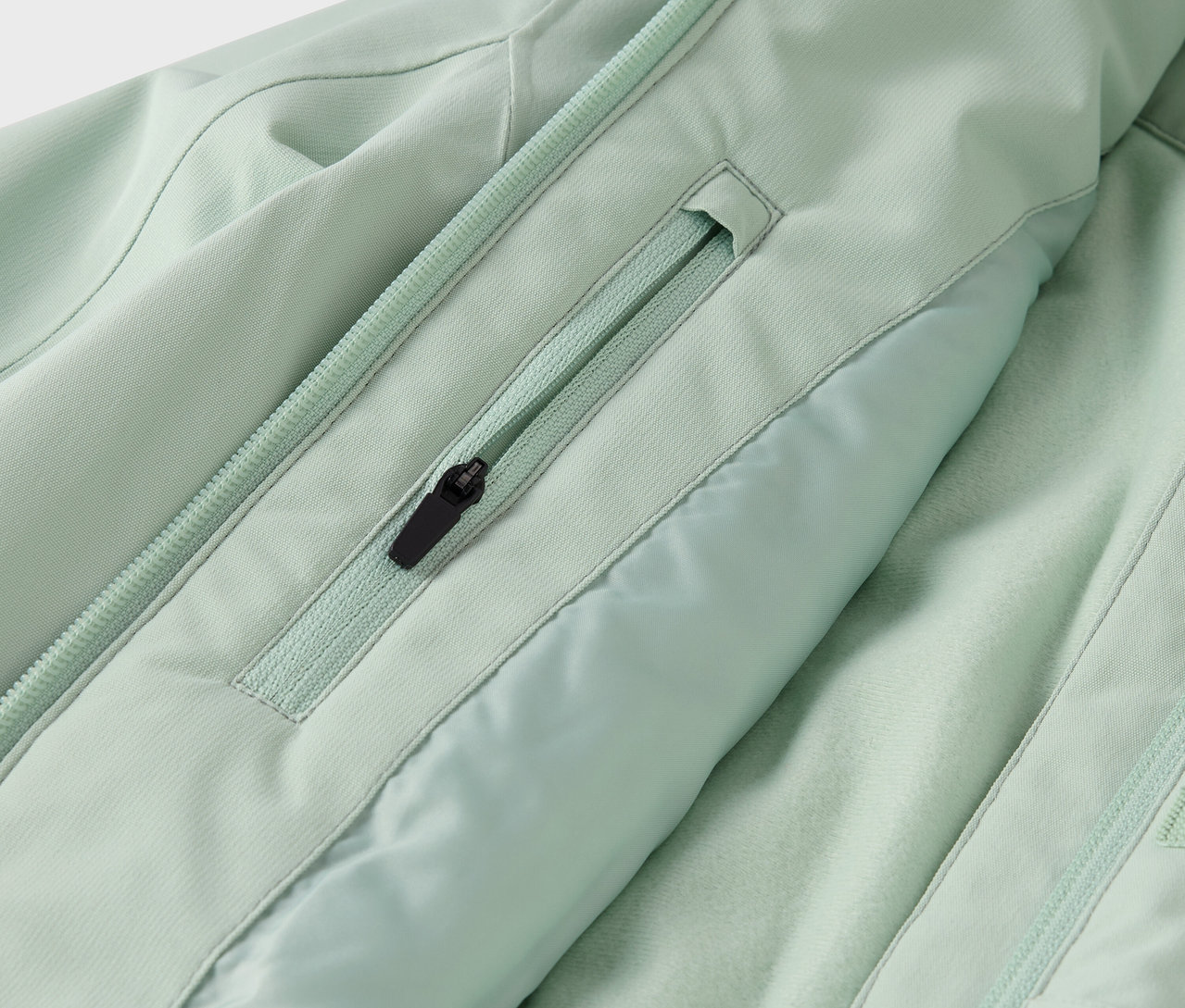 Szczegół jasnociemnozielonej kurtki z kieszenią na zamek błyskawiczny i zamkiem błyskawicznym pośrodku.