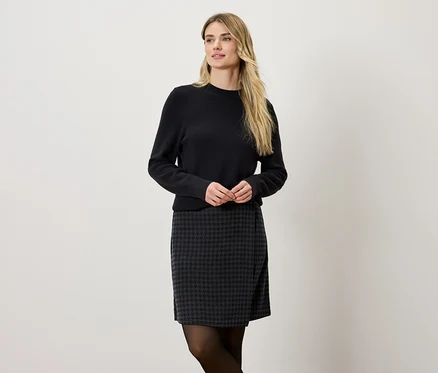 Blondynka ma na sobie czarny sweter i szaro-czarną spódnicę.